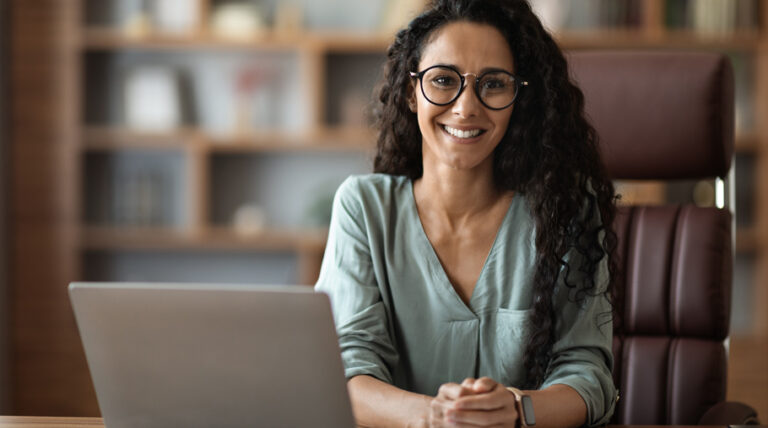 Donna sorridente in ufficio seduta al computer che guarda verso l’osservatore, simbolo di realizzazione professionale e passione trasformata in carriera grazie al coaching