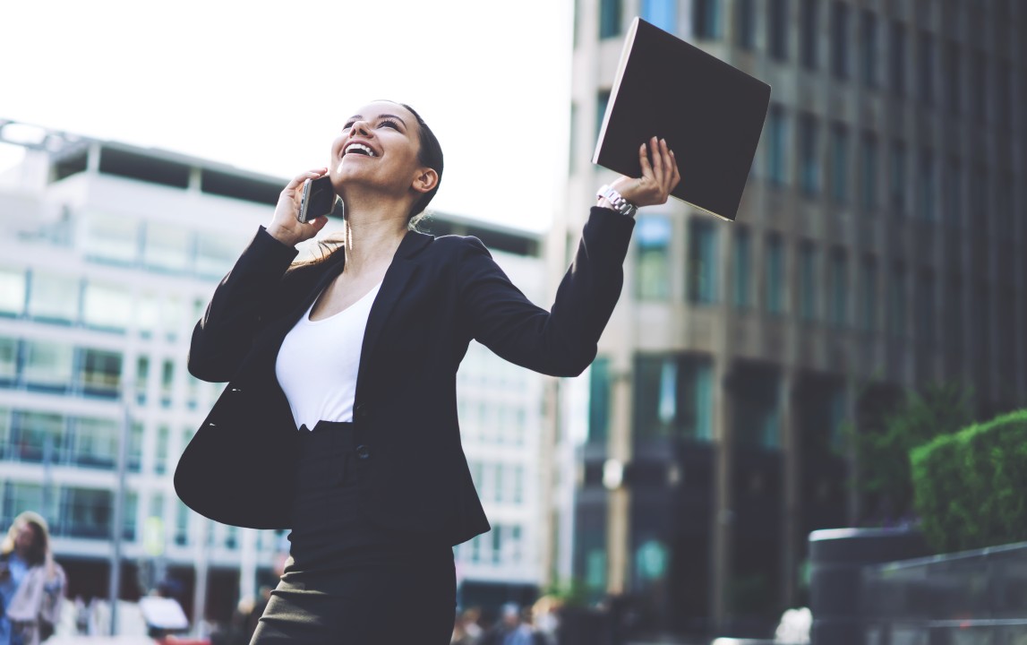 Donna sorridente in abbigliamento professionale che parla al telefono per strada, simbolo di successo, motivazione e raggiungimento degli obiettivi grazie al coaching